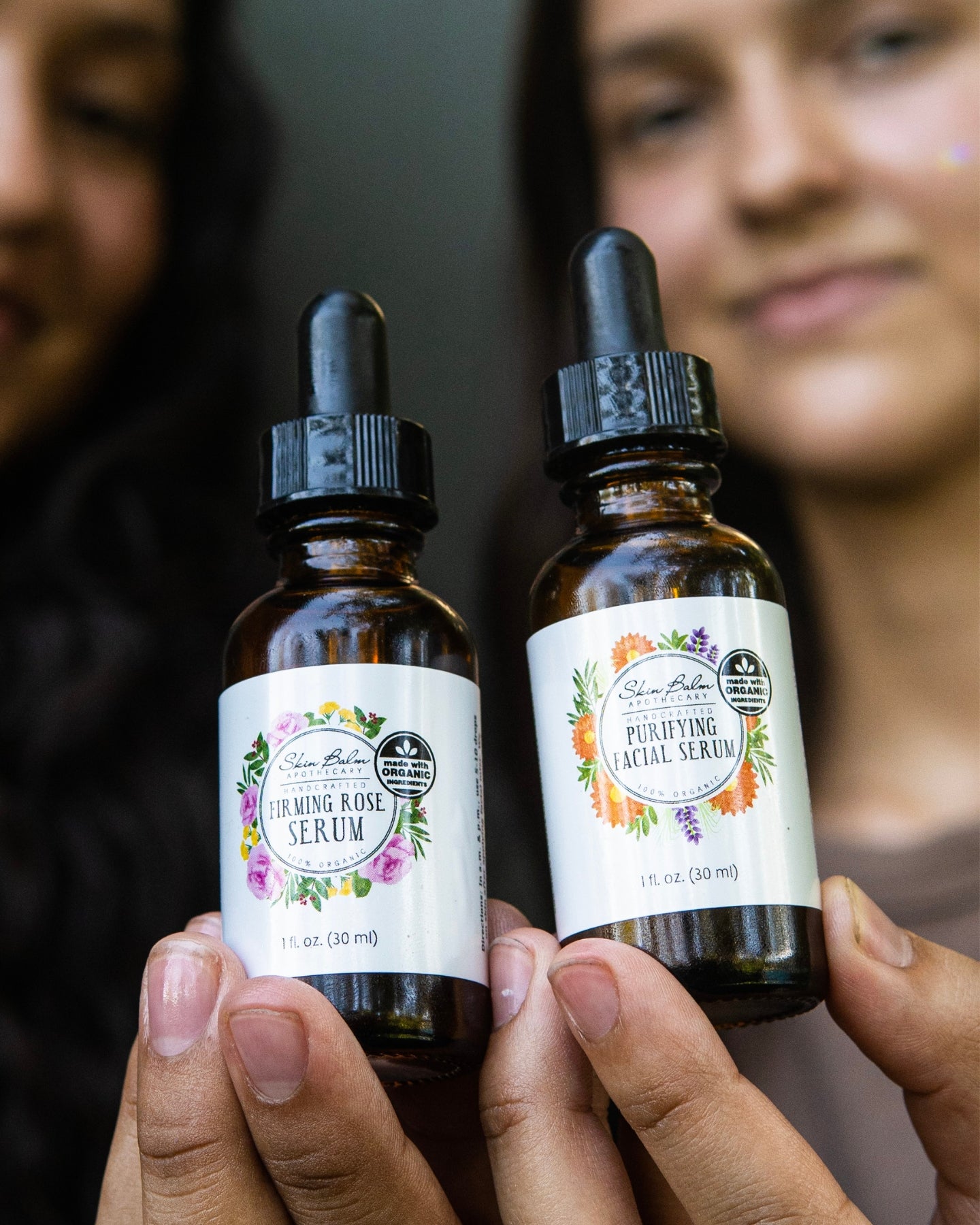 A close-up shot of the Firming Serum and the Purifying Serum being held side-by-side, with the two women holding the serums blurred in the background.