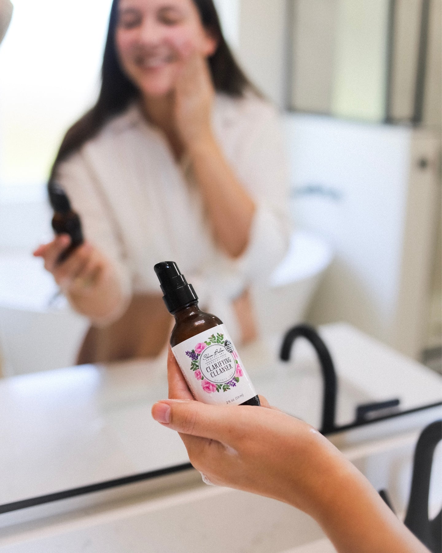 Clarifying Cleanser resting in a woman's hand focused in the foreground with a reflection of a woman rubbing the product onto her face blurred in the background.