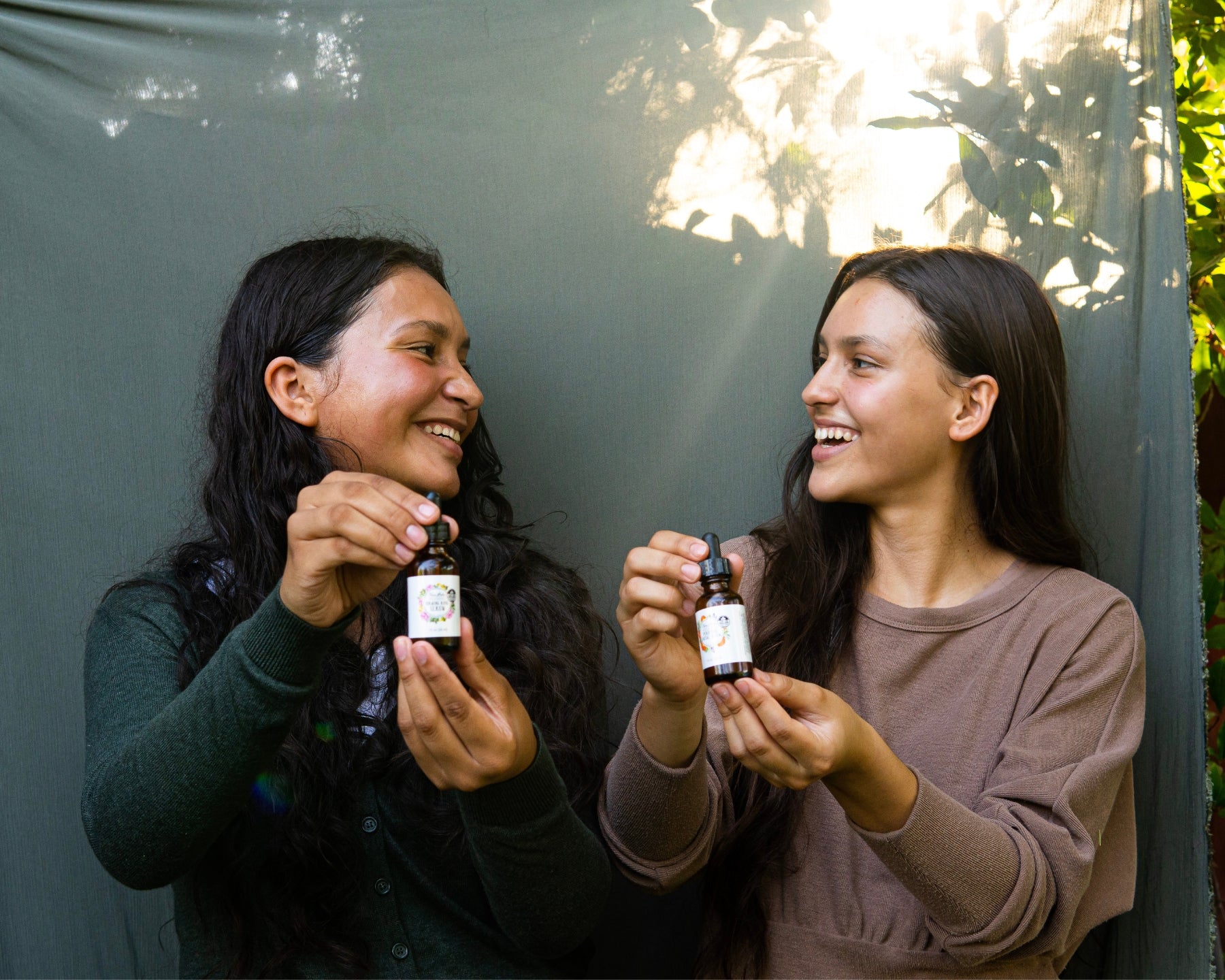 Two women look at each other and smile as they hold the Firming Serum and the Purifying Serum, with sunlight filtering through a sage green linen curtain in the background.